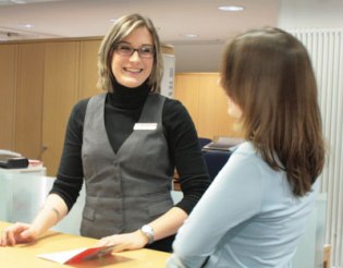 Ein Mädchen im Hosenanzug hinter einem Bankschalter.