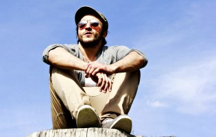 Junger Mann mit Sonnenbrille und Cap sitzt auf Baumstumpf und schaut in die Ferne