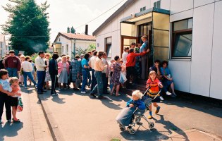 Menschenschlange vor Baracke, im Vordergrund Kind, das ein anderes im Kinderwagen schiebt