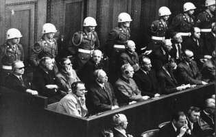 Reihe von Männern auf der Anklagebank mit Kopfhörern, dahinter Soldaten