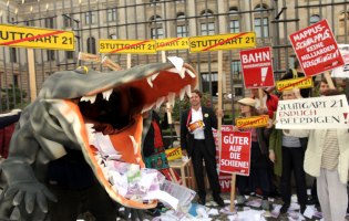 Plastikkrokodil mit aufgerissenem Maul, in dem Geldscheine liegen, im Hintergrund Menschen, die Plakate gegen Stuttgart 21 in die Höhen halten