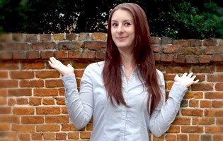 Alina steht vor einer Mauer und hebt fragend die Arme.