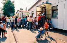 Menschenschlange vor Baracke, im Vordergrund Kind, das ein anderes im Kinderwagen schiebt