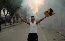 Junger Mann steht auf Straße, die Arme ausgebreitet, im Hintergrund Rauch/Tränengas